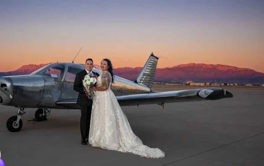 Newlyweds in front of a vintage airplane at sunset with the Sandia Mountains glowing pink